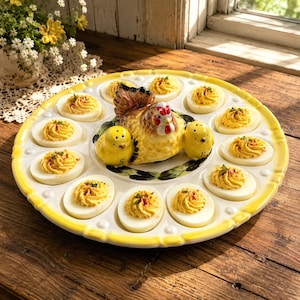 Puede incluir: Una bandeja redonda de cerámica para huevos rellenos con un borde festoneado amarillo. El centro presenta una gallina decorativa y dos pequeños saleros y pimenteros con forma de pollito. La bandeja contiene doce huevos duros rellenos.