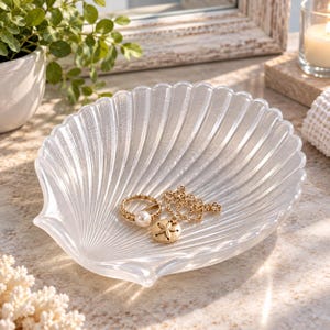 May include: A clear, translucent glass dish shaped like a scallop shell with a ribbed texture. Inside the dish rests a gold-toned ring featuring a white pearl and a gold-toned necklace with a small, circular sand dollar pendant charm.
