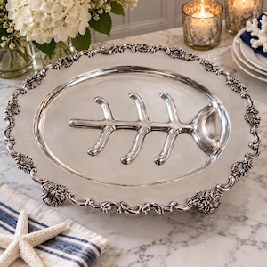 May include: An oval silver-toned serving platter featuring a raised fish skeleton design in the centre. The platter has a decorative scalloped edge with intricate shell-like detailing and rests on small feet, providing a polished metallic finish.