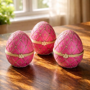 Puede incluir: Tres cajas decorativas rosas con forma de huevo y detalles dorados. Cada huevo tiene un patrón acolchado con detalles dorados y una banda dorada. Las cajas están sobre una superficie de madera.
