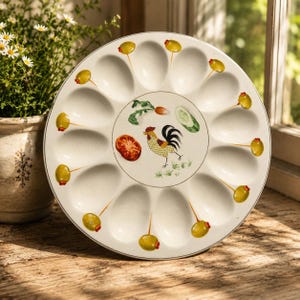 May include: A round, off-white ceramic egg serving platter featuring twelve moulded indentations arranged in a circle. The centre displays a colourful illustration of a rooster, a tomato slice, a cucumber slice, and a radish, with green olives on toothpicks painted between each egg cup.