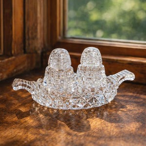 May include: A clear, cut-glass salt and pepper set. The set includes two shakers with a diamond pattern, resting on a matching tray with handles. The set is displayed on a wooden surface near a window.