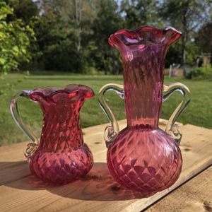 May include: Two cranberry-coloured glass vases with a diamond-quilted pattern and ruffled rims. The smaller vase features a single clear glass handle, while the larger, taller vase includes two clear glass handles on opposite sides of the neck.