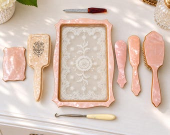 May include: A vanity set featuring a rectangular tray with a white lace inlay, two hairbrushes with light pink marbled handles, a small rectangular mirror, two small spatulas, a metal nail file, and a button hook with a cream-colored handle.