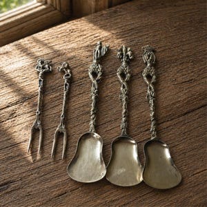 Puede incluir: Colección de utensilios de servir antiguos de plata, que incluye dos tenedores y tres cucharas. Los mangos están intrincadamente diseñados con figuras ornamentadas y patrones florales. Las cucharas tienen cuencos en forma de campana.