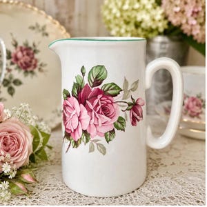 Könnte beinhalten: Weißer Keramikkrug mit grünem Rand und großem Henkel. Der Krug ist mit rosa und roten Rosen und grünen Blättern verziert. Ein passender Teller befindet sich im Hintergrund. Blumiges Thema.