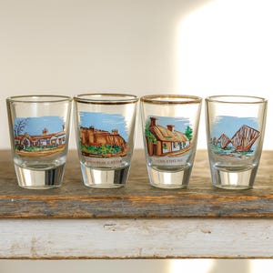 Op de afbeelding: Vier heldere shotglaasjes met gouden randen, elk met een andere schilderachtige illustratie. De afbeeldingen omvatten een huisje, een kasteel en andere architectonische ontwerpen. De glazen worden gepresenteerd op een rustiek houten oppervlak.
