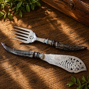 May include: A silver fork and a silver knife with dark brown textured antler handles. The fork features a four-tine design with floral engraving at the base. The knife blade is wide and decorated with intricate leaf and scrollwork patterns.