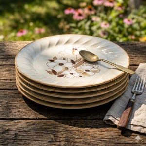 May include: A stack of five cream-colored ceramic plates with a scalloped edge and a brown floral pattern in the center. A brass-toned metal spoon rests on the top plate, and a silver-toned fork with a wooden handle lies on a beige cloth.
