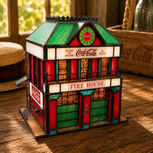 May include: A stained glass lamp shaped like a firehouse with red, green, and amber panels. It features Coca-Cola branding, including the text 'Delicious and Refreshing', 'Drink Coca-Cola in Bottles', and 'Fire House' on white glass panels.