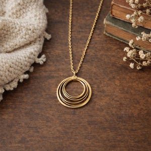 May include: A gold-colored necklace with a pendant of four nested circles hangs on a gold chain. The necklace rests on a dark wooden surface, with a cream-colored knitted scarf and vintage books in the background.