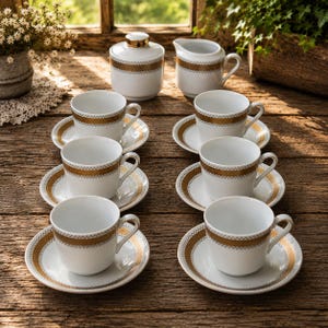 May include: A white porcelain tea set consisting of six cups and saucers, a lidded sugar bowl, and a creamer pitcher. Each piece features a wide, textured gold band with a repeating geometric pattern around the center and the saucer rims.