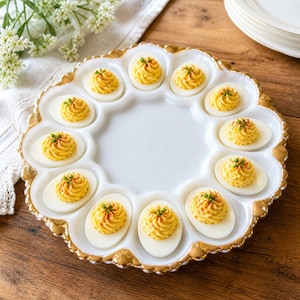 Könnte beinhalten: Eine weiße Servierplatte für gefüllte Eier mit goldenem Rand, die zwölf gefüllte Eier enthält. Jede Eihälfte ist mit gelber Füllung gefüllt und mit Paprika und Schnittlauch garniert. Die Platte steht auf einer Holzoberfläche.