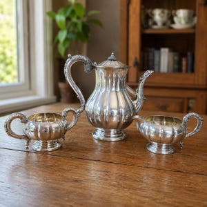 May include: A silver tea set comprising a teapot, a sugar bowl and a creamer. Each piece features a polished metallic finish, vertical fluted detailing, and ornate, curved handles with intricate embossed patterns on the edges.