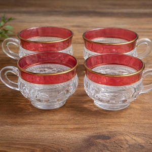 May include: A set of four clear glass teacups featuring a wide, deep red band around the upper rim, accented with a thin gold trim. The lower portion of each cup displays a textured, pressed glass pattern with a rounded base and a curved handle.