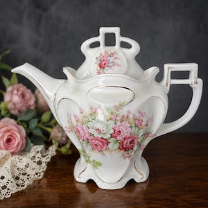 Op de afbeelding: Een witte porseleinen theepot met een bloemmotief van roze en witte rozen. De theepot heeft gouden details en een uniek handvat en deksel. De achtergrond is donkergrijs en de theepot staat op een houten oppervlak.