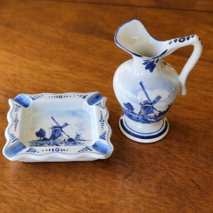 Może przedstawiać: Biało-niebieski ceramiczny zestaw dzbanek i popielniczka. Dzbanek ma zakrzywioną wylewkę i uchwyt. Oba elementy zdobi niebieski krajobraz z wiatrakami. Popielniczka jest prostokątna z falistą krawędzią.