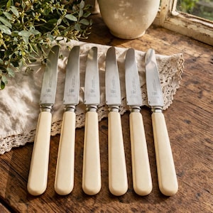 May include: Six vintage butter knives with cream-colored handles and silver blades are arranged on a wooden surface. The knives are placed on a lace doily, with greenery and a ceramic bowl in the background. The handles have a curved shape.