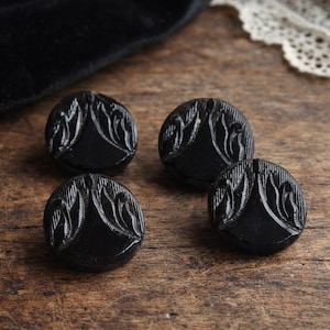 Puede incluir: Cuatro botones negros redondos con un patrón floral o de hojas tallado en relieve sobre la superficie. Cada botón tiene un acabado liso y brillante, con una forma circular y un diseño grabado distintivo que irradia desde el centro hacia los bordes.
