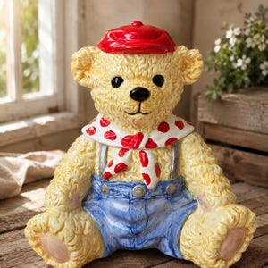Pode incluir: Uma estatueta de urso de peluche em cerâmica com pelo amarelo texturizado, usando um boné vermelho brilhante, um lenço branco com bolinhas vermelhas e jardineiras azuis com botões prateados. O urso tem olhos e nariz pretos, sentado de frente.