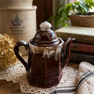 May include: A brown ceramic teapot with a white and brown glazed rim and lid. The teapot has a geometric design and a curved handle. A vintage stoneware crock is in the background. The teapot is sitting on a lace doily.