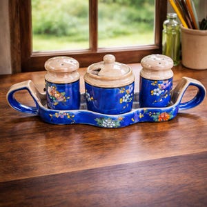 Puede incluir: Un juego de condimentos de cerámica azul con una bandeja. El juego incluye dos saleros y un tarro con tapa, todos con diseños florales. La bandeja y las tapas de los recipientes son beige.