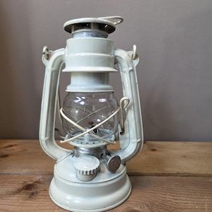 May include: A cream-colored metal hurricane lantern featuring a clear glass globe protected by a wire cage. The lantern includes a top handle, a side adjustment knob, and a fuel cap on the base, showcasing a classic oil lamp design.