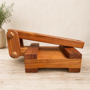 Puede incluir: Una prensa de tortillas de madera con un brazo de palanca. La prensa está hecha de madera marrón claro y tiene una base rectangular con un brazo articulado para prensar tortillas. El diseño es simple y rústico, adecuado para hacer tortillas caseras.
