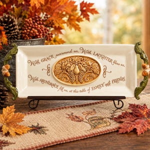 Puede incluir: Una bandeja de servicio rectangular de cerámica color crema con asas en forma de hoja verde y detalles de bellotas. El centro muestra un relieve ovalado de una pera y uvas, rodeado por el texto: With GRACE surround us, With LAUGHTER bless us, With ABUNDANCE fill us, at this table of FAMILY and FRIENDS.