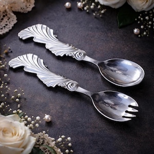 May include: Two silver serving spoons with ornate, stylized handles. One spoon has a traditional bowl, while the other has a three-pronged fork. The handles feature a flowing, leaf-like design with a central swirl. The spoons are set against a dark background.