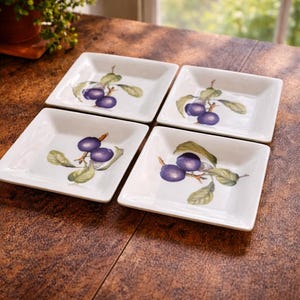 May include: Four square white ceramic dishes with a plum branch design. Each dish features a cluster of purple plums with green leaves and brown stems. The dishes are arranged on a dark brown wooden surface.