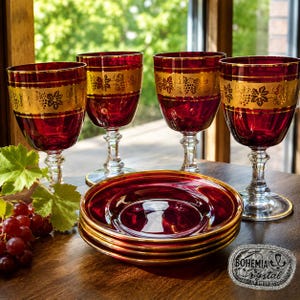 Peut inclure: Ensemble de quatre verres à vin rouges et des assiettes assorties, avec des bordures dorées et des motifs de grappes de raisin. Les verres et les assiettes en cristal sont présentés sur une table en bois. Le logo Bohemia Crystal est visible.