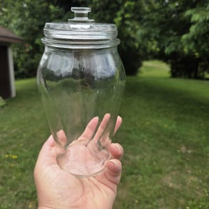 Puede incluir: Un frasco de vidrio transparente con cuerpo cónico y una tapa de vidrio a juego que presenta un pequeño mango en forma de perilla en la parte superior. El frasco tiene una boca ancha y una superficie lisa y transparente para el almacenaje.