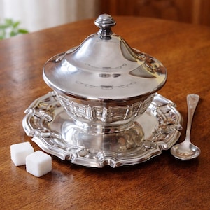 May include: A silver-plated sugar bowl with a lid, resting on a matching tray, and a small spoon. Two white sugar cubes sit on the wooden surface. The bowl has a decorative, ornate design.