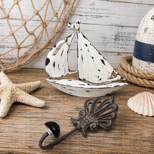 Pode incluir: Arranjo decorativo com tema náutico com um veleiro branco envelhecido com detalhes pretos, uma estrela do mar, uma concha e um gancho decorativo. O veleiro tem cerca de 20 cm de comprimento. Os objetos estão dispostos sobre uma superfície de madeira.