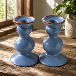 May include: A pair of blue ceramic candle holders with a rounded, stacked bulbous silhouette. Each holder features a wide circular base, a narrow neck, and a flared top rim accented with a thin brown edge and small light blue rectangular details.
