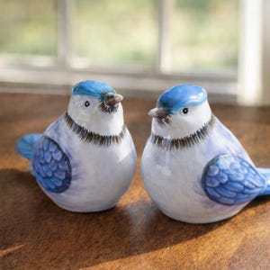May include: Two ceramic blue jay ornaments. Each bird features a blue head and wings, a white body, and a black collar. The ornaments are displayed on a wooden surface near a window.