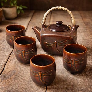 Puede incluir: Un juego de té de cerámica marrón con una tetera con asa trenzada y tazas a juego. Cada pieza presenta un diseño de libélula. El juego se exhibe sobre una superficie de madera rústica, creando una estética cálida y acogedora.