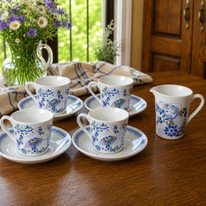 May include: A set of four white ceramic teacups and matching saucers alongside a matching creamer pitcher. Each piece features a blue and green graphic illustration of a stylized figure surrounded by floral patterns and intricate decorative borders.