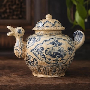 May include: A cream-coloured ceramic teapot with a duck-shaped spout and handle. It features blue floral patterns and a cracked glaze. The lid has a small knob. The teapot is on a wooden surface.