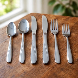 Puede incluir: Un juego de seis cubiertos de acero inoxidable, que incluye dos cucharas, dos cuchillos y dos tenedores, dispuestos sobre una superficie de madera. La cubertería tiene un diseño sencillo y elegante con mangos curvos.