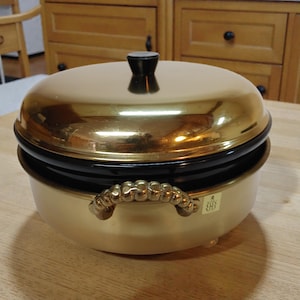 Może przedstawiać: Złoty garnek z pokrywką, czarnym paskiem i uchwytem. Garnek ma zaokrąglony kształt i czarny guzik na pokrywce. Uchwyt jest ozdobny i złoty. Garnek stoi na drewnianej powierzchni.