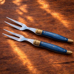 May include: Two stainless steel carving forks with blue wooden handles. The handles have brass-coloured metal bands and rivets. The forks are on a wooden surface.