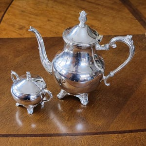 Op de afbeelding: Een glanzende zilveren theeset, bestaande uit een theepot en een suikerpot, staat op een houten oppervlak. De theepot heeft een gebogen tuit en een sierlijke handgreep. De suikerpot heeft een deksel en kleine handgrepen. De set weerspiegelt de omgeving.