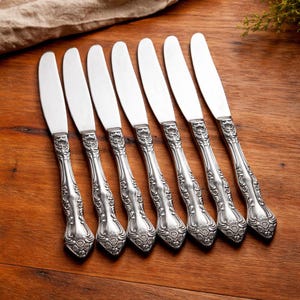 Könnte beinhalten: Ein Set aus sieben silbernen Buttermessern mit verzierten, floral gemusterten Griffen. Die Messer sind auf einer Holzoberfläche angeordnet und zeigen ihre polierten Klingen und das detaillierte Design. Die Griffe weisen eine klassische, Vintage-Ästhetik auf.