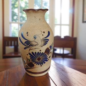 May include: A beige ceramic vase with a textured surface and a ruffled rim. The vase features a hand-painted design of a blue bird and a blue flower. The base and rim are a dark brown colour. The vase is on a wooden table.