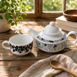 Könnte beinhalten: Eine weiße Keramik-Teekanne und eine passende Teetasse mit einem schwarzen Blumenmuster und vertikalen Linien. Die Teekanne hat einen abgerundeten Deckel und einen geschwungenen Griff, während die Tasse eine breite Schalenform aufweist.