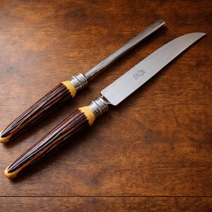 May include: A knife and sharpening steel set. The knife has a curved blade and a handle with brown and cream tones. The sharpening steel has a similar handle design. Both pieces have silver-colored metal accents.