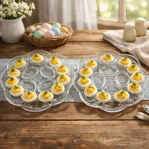 Puede incluir: Dos fuentes de vidrio transparente para servir huevos rellenos con un diseño floral, cada una con 12 huevos rellenos. Una cesta de huevos de colores pastel y un jarrón de flores blancas están en el fondo, sobre una mesa de madera.