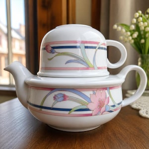 Könnte beinhalten: Weißes Keramik-Teeservice mit floralem Design. Die Teekanne hat einen Ausguss und einen Henkel, und die Tasse sitzt obenauf. Das Design umfasst rosa, blaue und grüne Blumen- und Streifenakzente. Das Set steht auf einer Holzoberfläche.